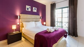 Un dormitorio con cama, dos lámparas de pared, una mesita de noche, una colcha morada y dos cuadros enmarcados con motivos florales.