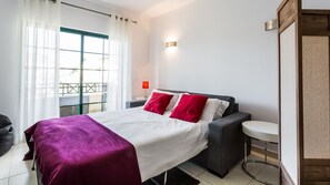 Quarto de hotel moderno com uma cama grande, uma mesinha de cabeceira, uma mesa redonda pequena e vista para a varanda externa.