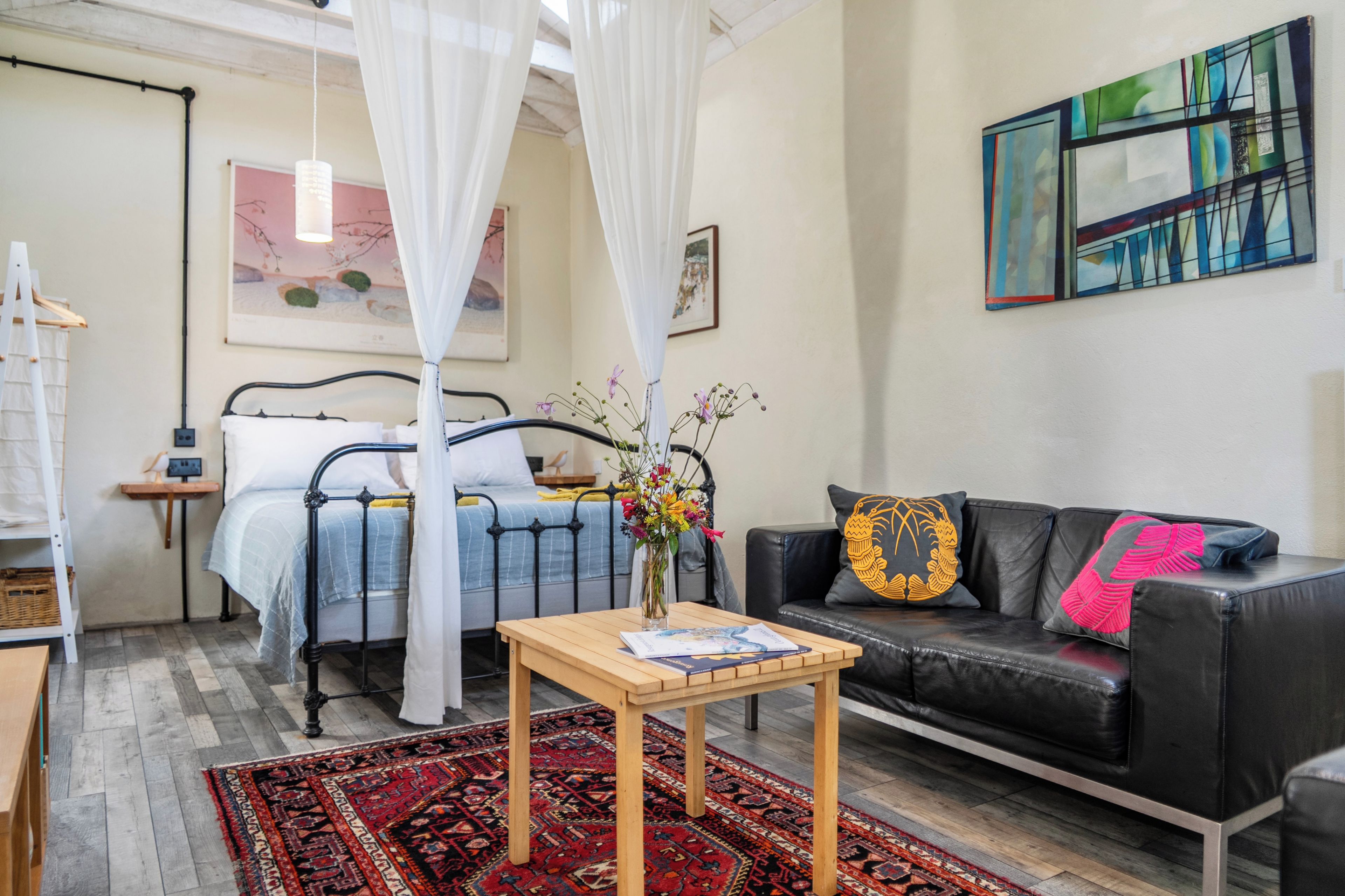 A bedroom with a bed, a bedside table with a book, a black leather sofa, a colorful rug, and a painting on the wall.