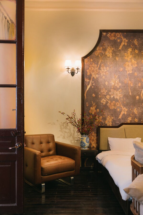 Habitación de hotel con una butaca de cuero marrón, una cama con ropa blanca, una lámpara de pared y un jarrón decorativo con flores.