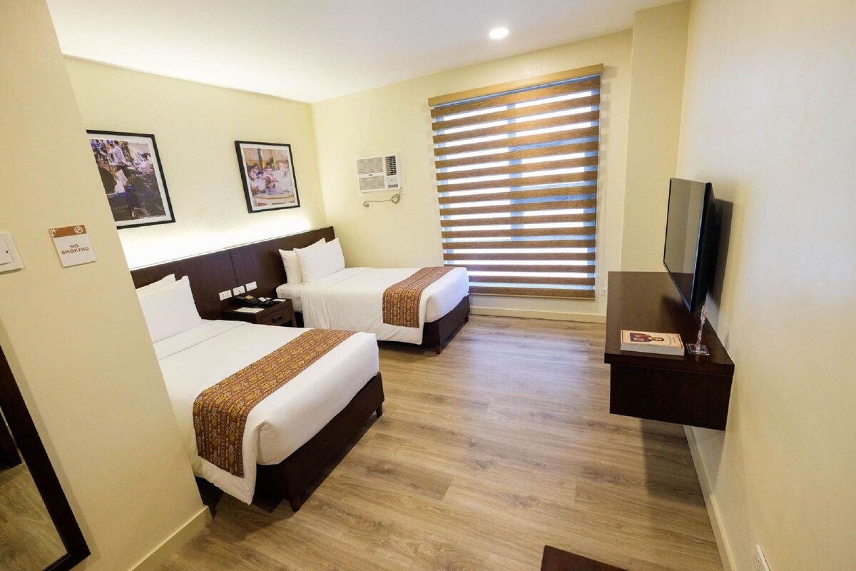 A hotel room with two beds, a TV, and a window with blinds.