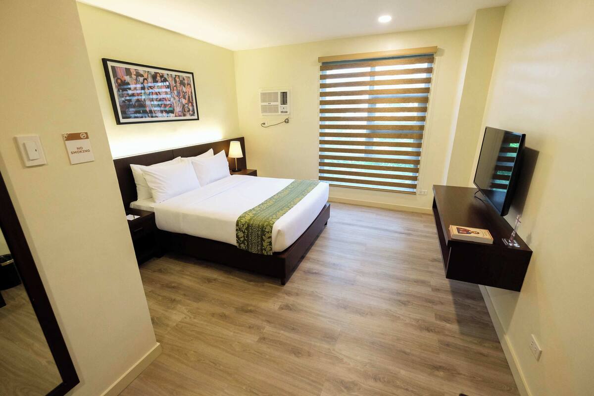 A hotel room with a bed, a TV, a window with blinds, a picture on the wall, and a sign indicating no smoking.