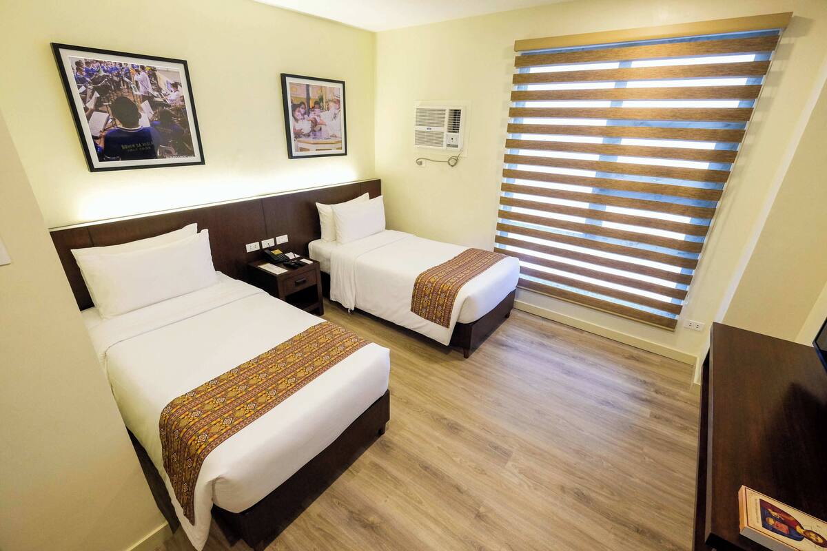 A hotel room with two beds, a wooden floor, a large window with blinds, and a wall-mounted air conditioner.
