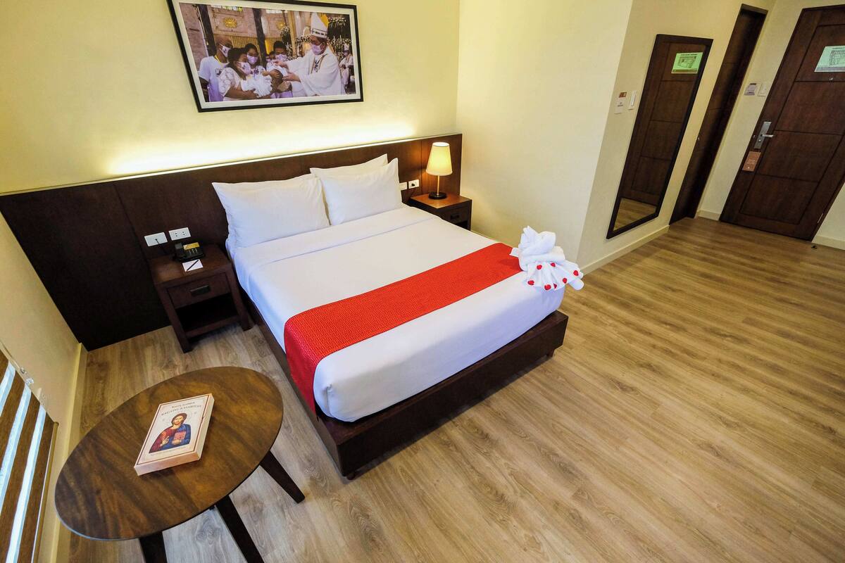 A hotel room with a neatly made bed, a bedside table with a book, a lamp, and a framed photo on the wall.