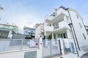 A white multi-story residential building with balconies, a gated entrance, and a clear sky.