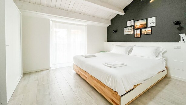 Un dormitorio moderno con una cama de madera, ropa de cama blanca, un ventanal grande y una pared oscura con cuadros enmarcados.