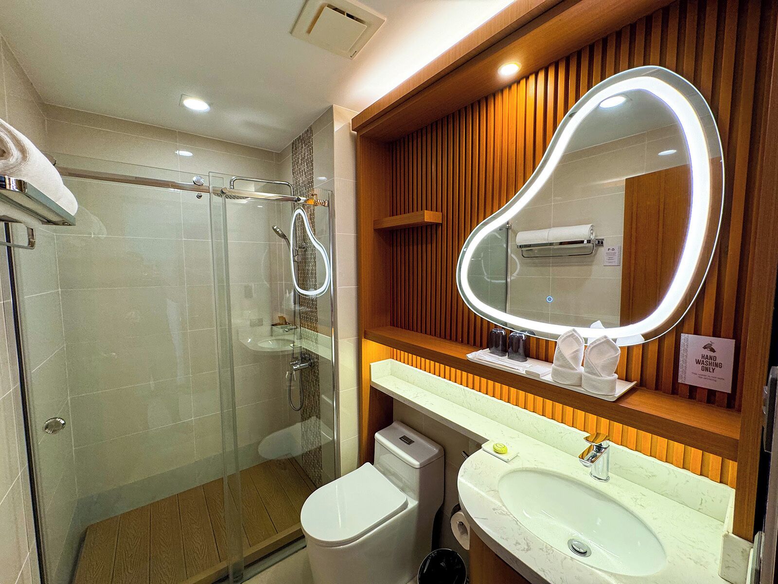 A modern bathroom with a glass-enclosed shower, a white toilet, a sink with a mirror, and wooden paneling on the wall.