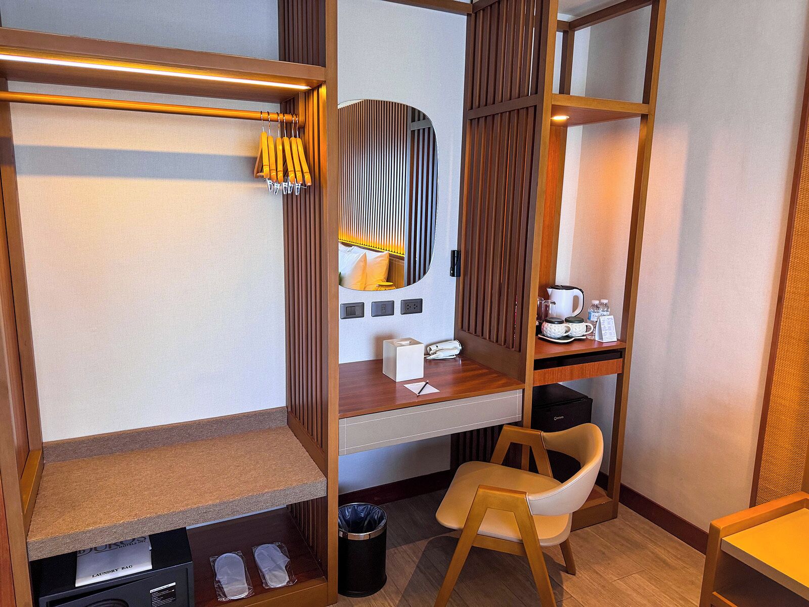 A compact hotel room with a wooden desk, a chair, a small trash bin, and a shelf with toiletries.