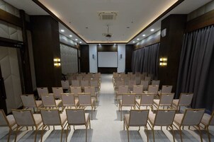 Un salón de conferencias con filas de sillas, una pantalla de proyector y una zona de escenario.