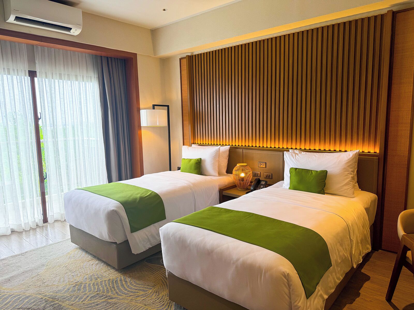 A hotel room with two beds, a large window with curtains, a wall-mounted lamp, and a wooden panel design on the wall.