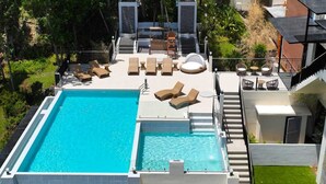 Una piscina en la azotea con sillones de descanso y una cama solárium, rodeada de follaje y un edificio moderno.