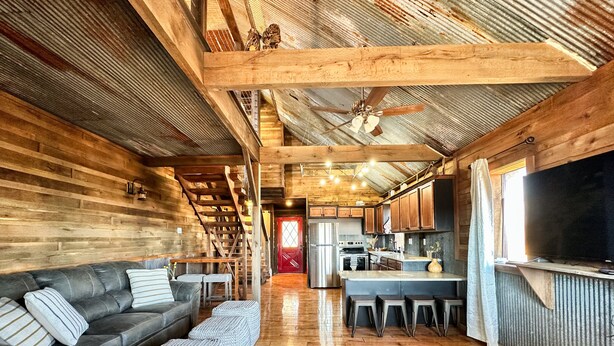 A rustic cabin interior with a wooden staircase, a sofa, ottomans, a kitchen area, and a ceiling fan. - Private, rustic country home with hot tub, movie theater and a view (Pineville)
