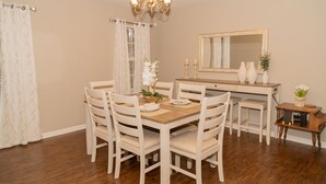 A dining area with a wooden table and white chairs, a chandelier, a mirror, and a small side table with decorative items. - Beautiful spacious 4 bedroom house , sleeps 10+ (Raleigh)