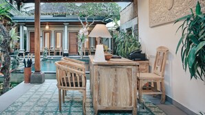 A wooden table with chairs, a lamp, and a decorative wall piece in a covered outdoor area with a pool and tropical plants. - Sitala Suite (Ubud)
