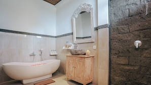 A bathroom with a freestanding bathtub, a wooden cabinet, a large mirror, and a stone wall. - Sitala Suite (Ubud)