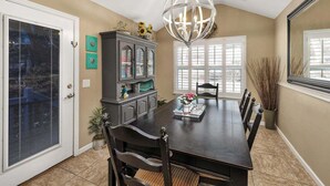 Comedor con mesa de madera oscura, sillas, candelabro, aparador con objetos decorativos y ventana con persianas.