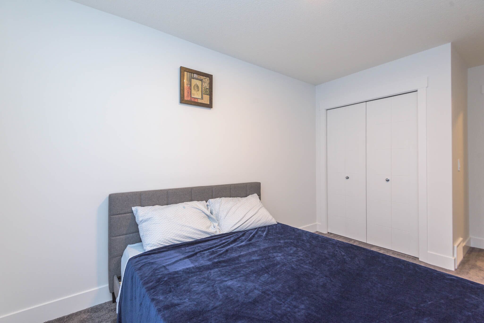 A bedroom with a bed, a grey headboard, a blue bedspread, a white wall, and a white door with a handle.