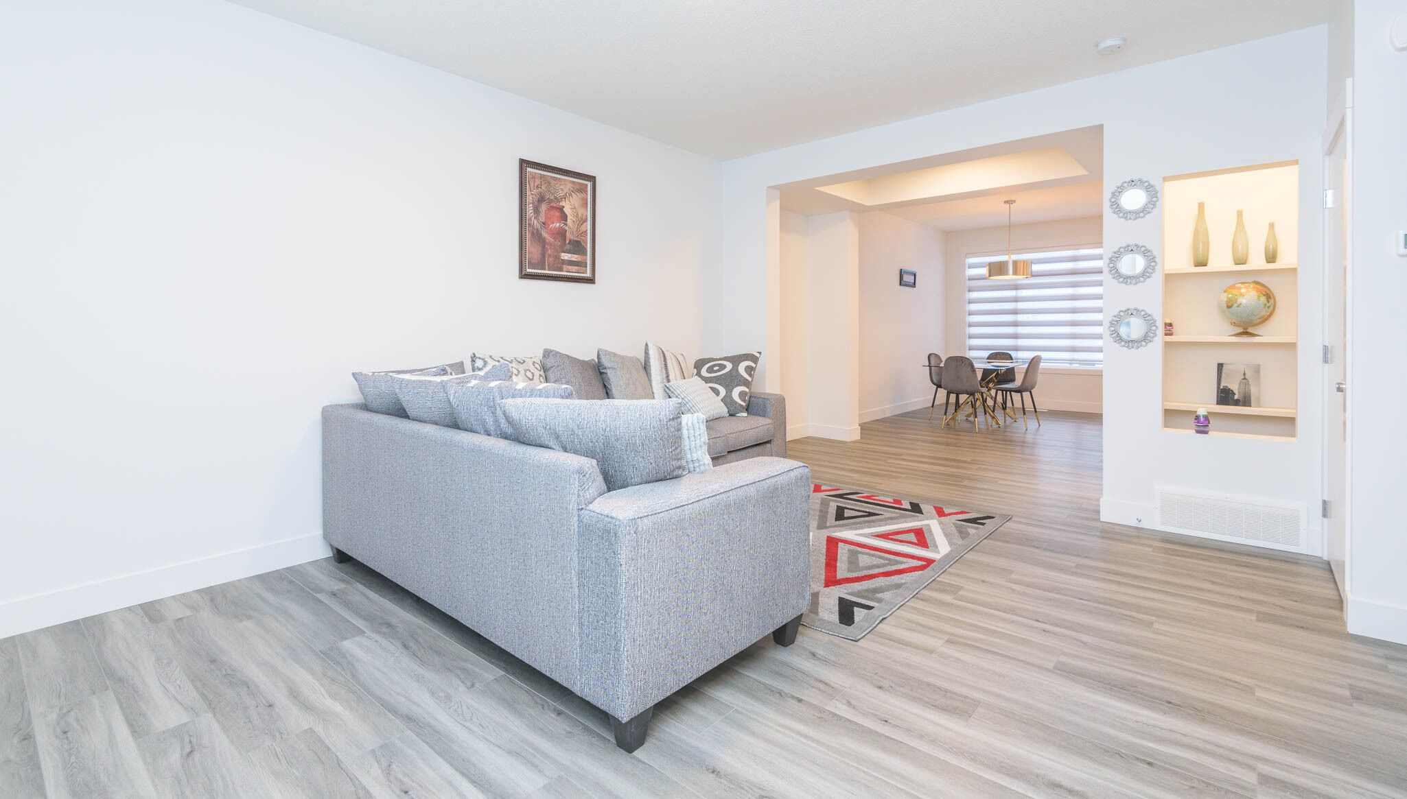 Een moderne woonkamer met een grijze bank, een eethoek met een tafel en stoelen, en een gang die naar een andere kamer leidt.