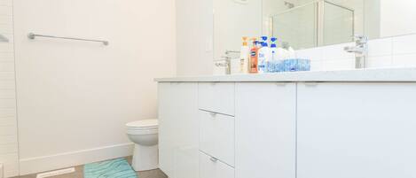 Ein Badezimmer mit Toilette, Dusche und Waschtisch mit Spiegel. Handtücher liegen auf dem Boden und Pflegeprodukte stehen auf dem Waschtisch.