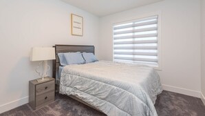 A bedroom with a bed, a nightstand, a lamp, a window with blinds, and a motivational poster on the wall. - Luxury and Comfort Combined. (Edmonton)