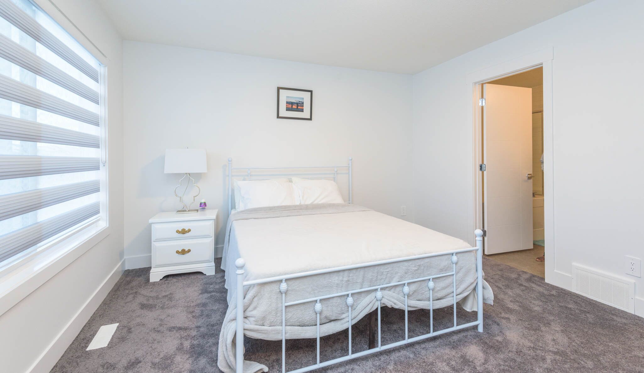 Un dormitorio con una estructura de cama metálica blanca, una mesita de noche con cajones, una lámpara y un cuadro en la pared.