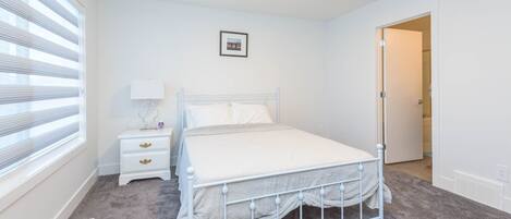 Un dormitorio con una estructura de cama metálica blanca, una mesita de noche con cajones, una lámpara y un cuadro en la pared.
