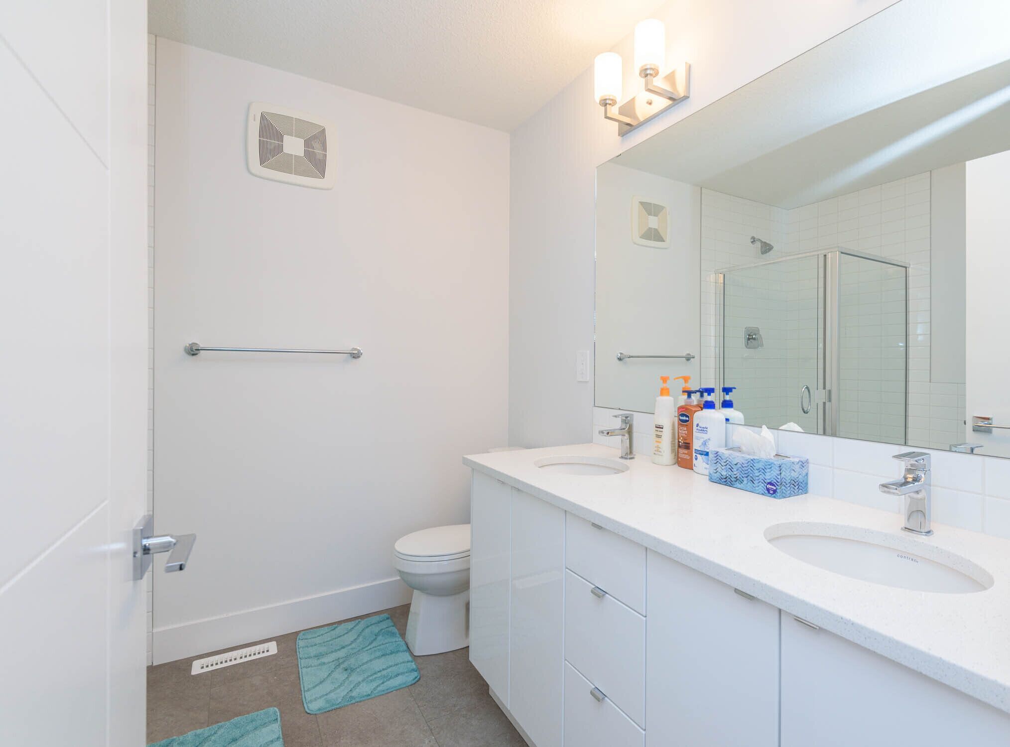 A modern bathroom with a white sink, vanity, and a large mirror. There is a towel rack, a toilet, and a shower area visible.