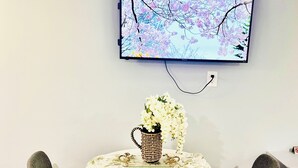 A dining area with a round table, two gray chairs, a woven basket with white flowers, and a flat-screen TV mounted on the wall displaying a cherry blossom tree.
