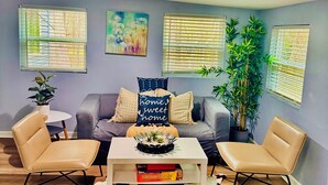 A living room with a grey sofa, two beige armchairs, a white coffee table, and a cowhide rug. There are two windows with blinds, a painting on the wall, and a potted plant.