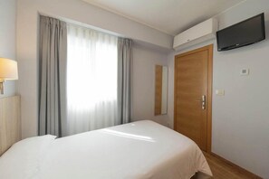 A hotel room with a bed, a TV mounted on the wall, a wooden door, and a window with curtains. - Hotel Panton (Vigo)