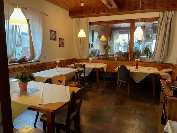 A cozy dining area with wooden tables and chairs, large windows with curtains, and hanging pendant lights.