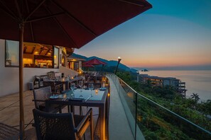 Uma área de refeições ao ar livre com mesas e cadeiras, com vista panorâmica para o mar e colinas.
