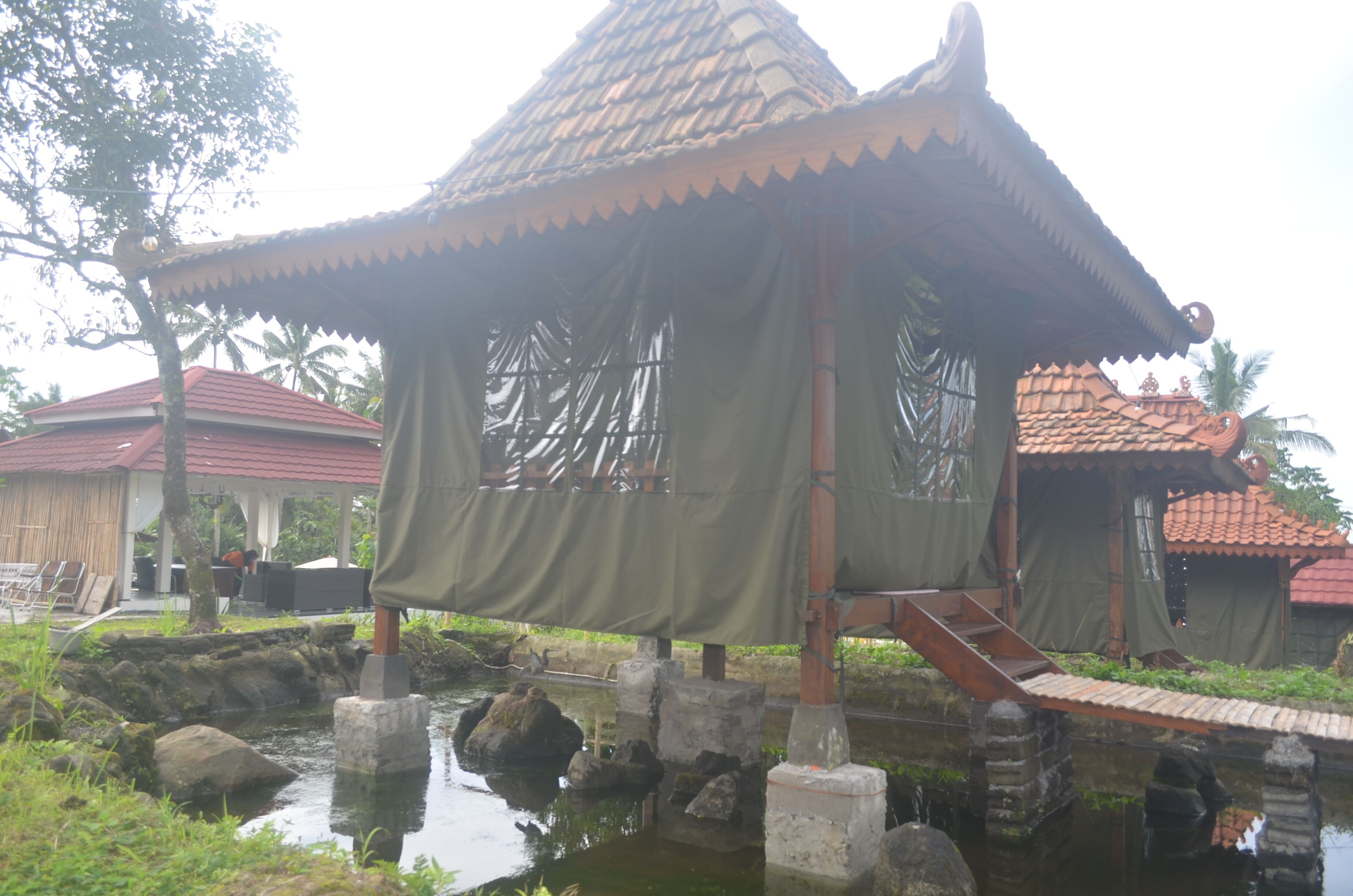 Een klassiek paviljoen met een betegeld dak, ondersteund door stenen zuilen, boven een kleine waterpartij.