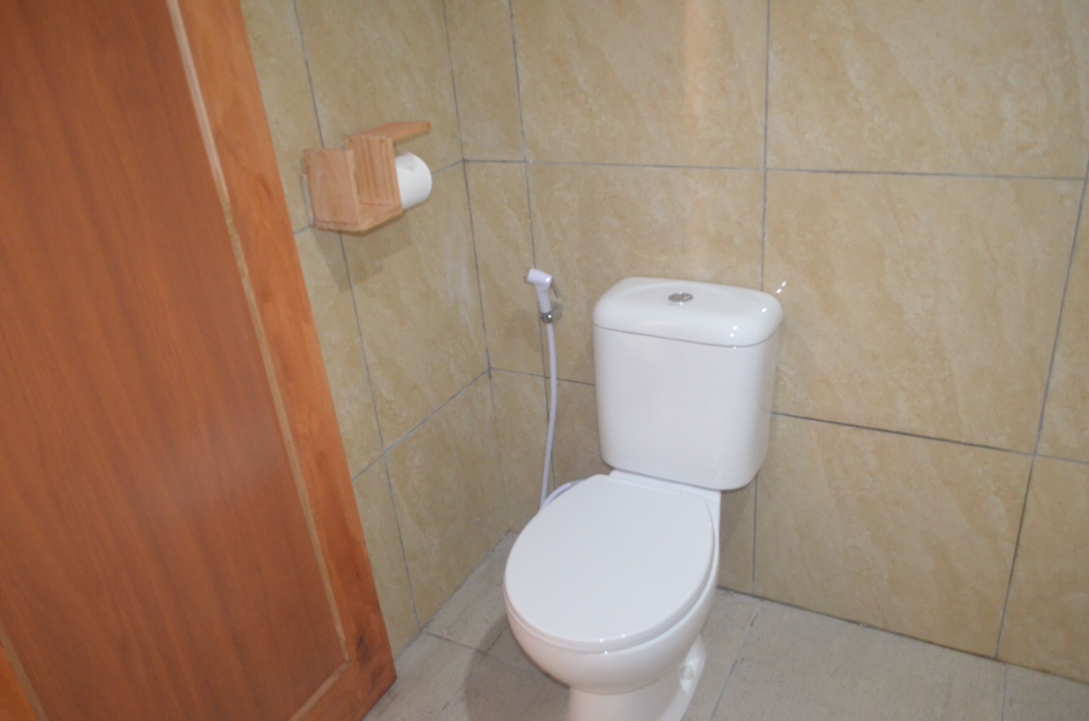 Un baño con inodoro, un estante de madera con dispensador de papel higiénico y paredes cubiertas de baldosas beige.
