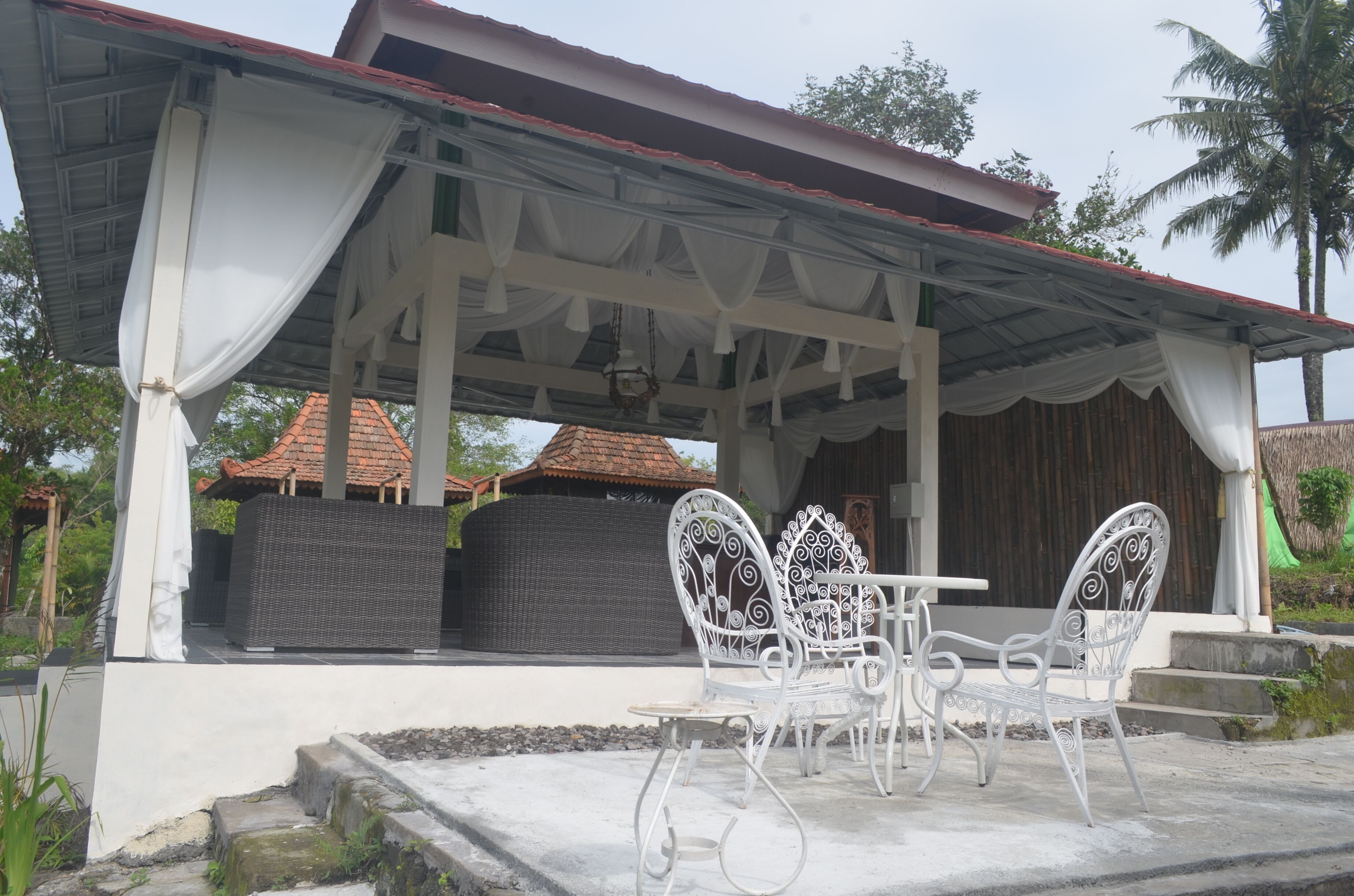 A covered outdoor seating area with white metal chairs and a table, a covered structure with a red roof, and a wicker sofa.