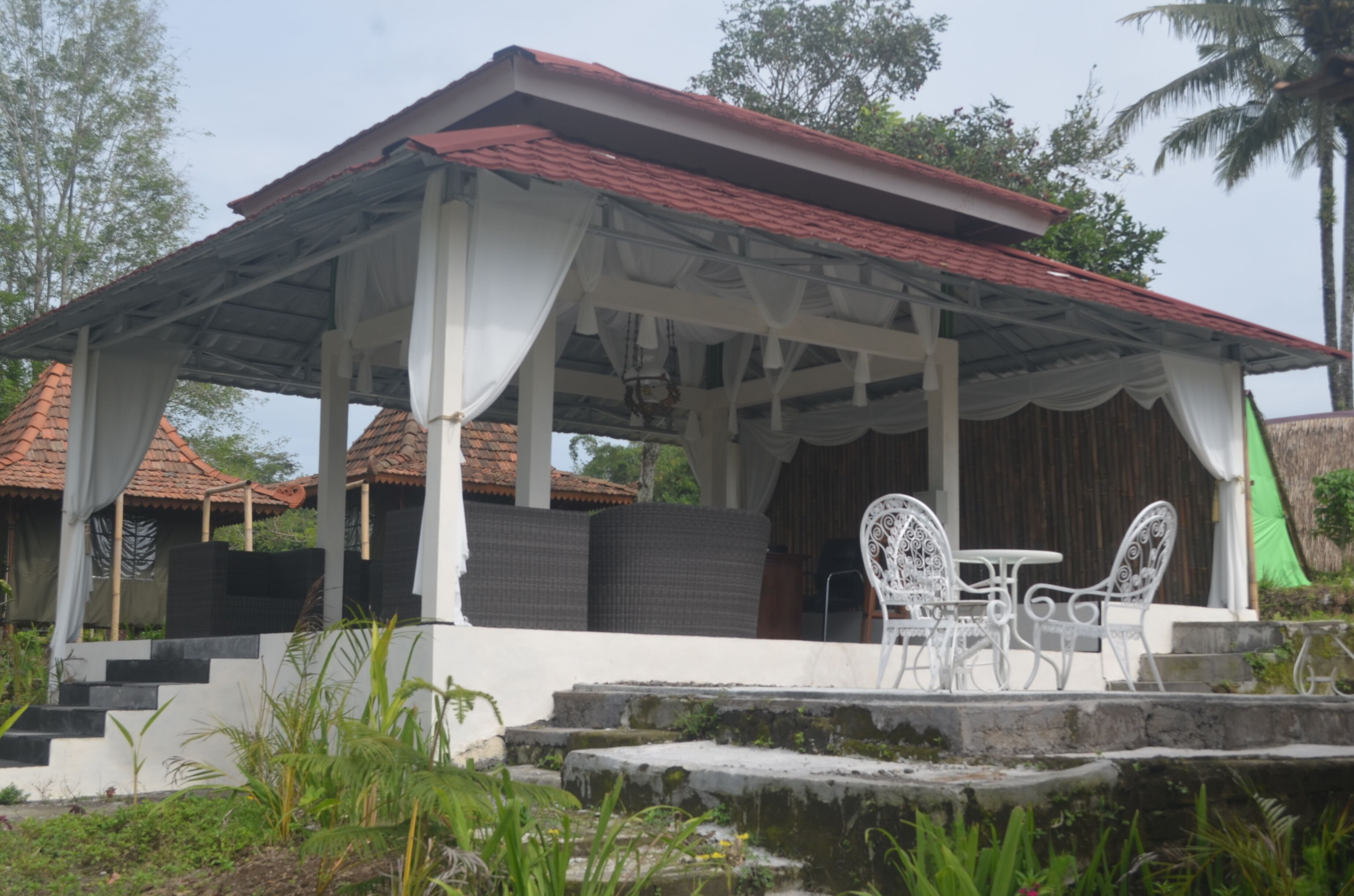A covered outdoor seating area with white metal chairs and a table, surrounded by steps and greenery.