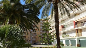 A multi-story hotel with balconies, a well-maintained garden with palm trees and shrubs, and a clear blue sky. - Cye 5 Rentalmar (Vila-Seca)