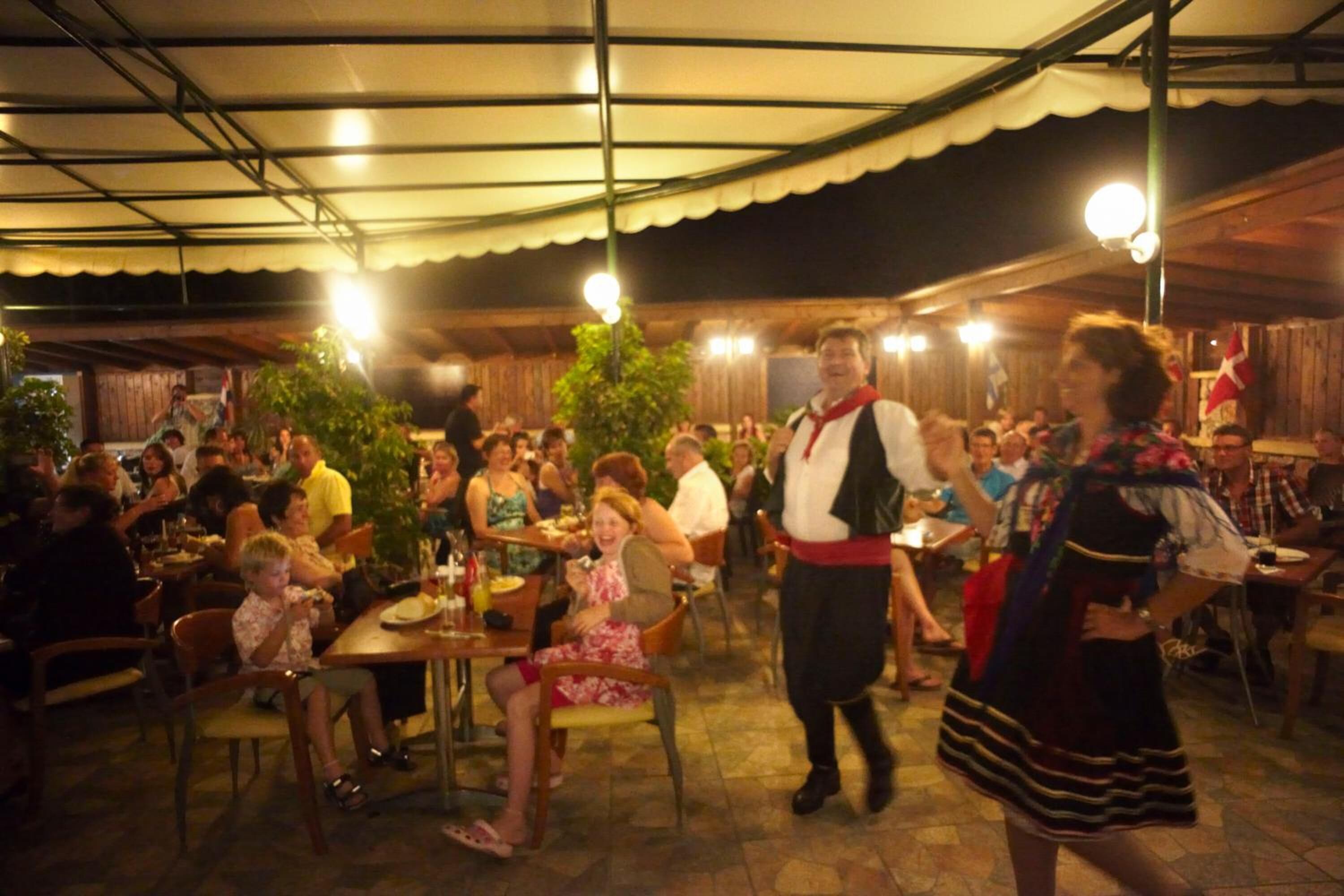A covered outdoor dining area with tables and chairs, surrounded by a crowd of people.