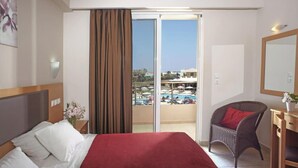 A hotel room with a bed, a chair, a desk, and a view of a pool and buildings. - Emerald Apartments (Rhodes)