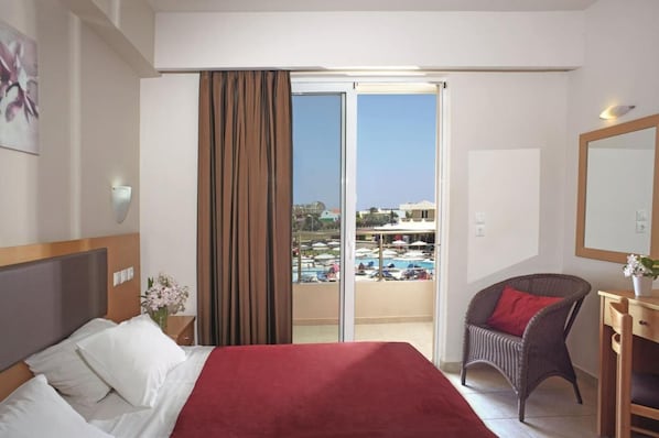 A hotel room with a bed, a chair, a desk, and a view of a pool and buildings.