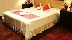 Una cama bien hecha con una colcha blanca y almohadas rojas, un cabecero de madera y una mesita de noche con una lámpara y flores.