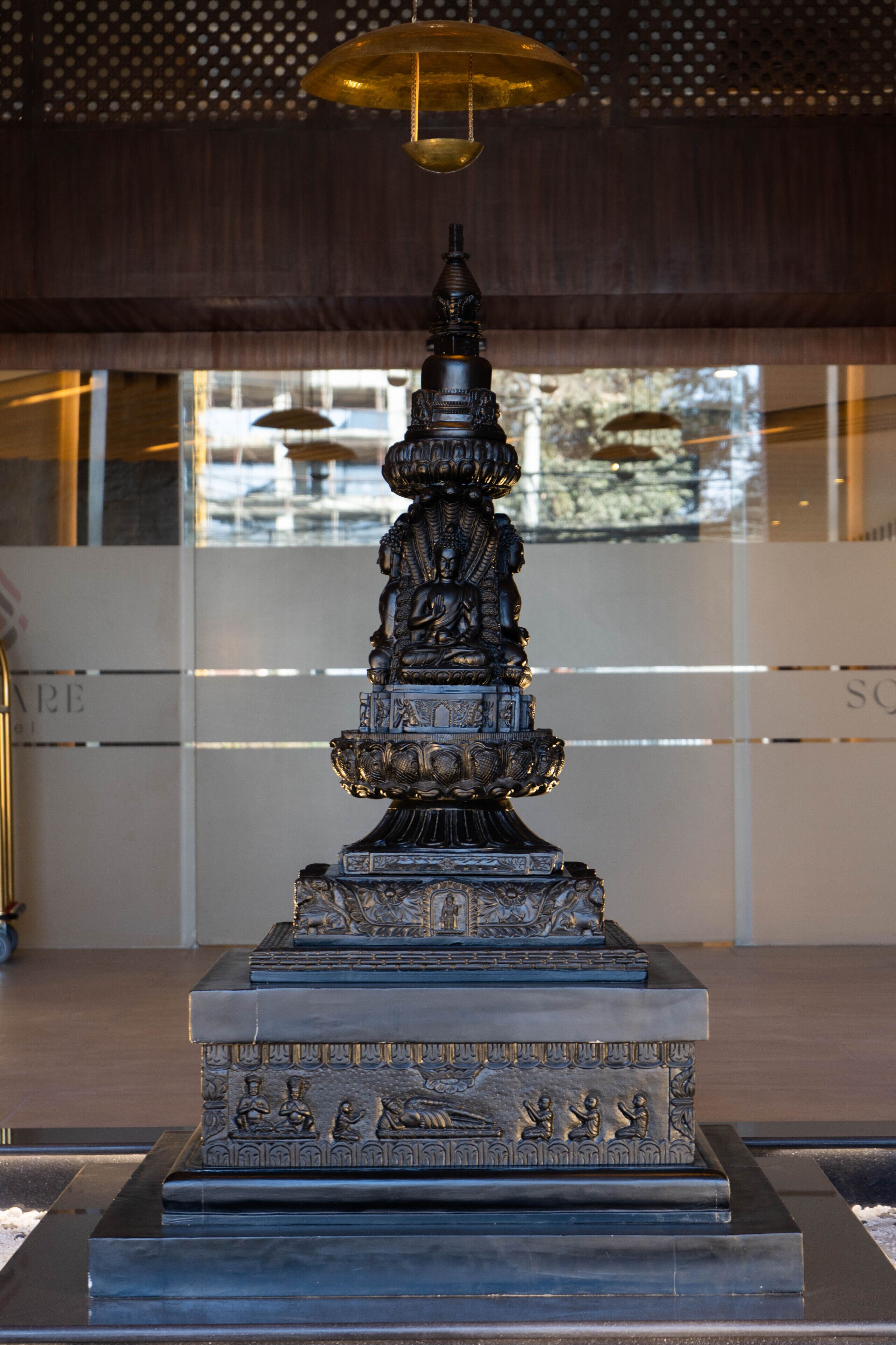 Una estatua de bronce con tallas detalladas sobre un pedestal, ubicada en un interior moderno con paredes de vidrio y un techo con diseño perforado.