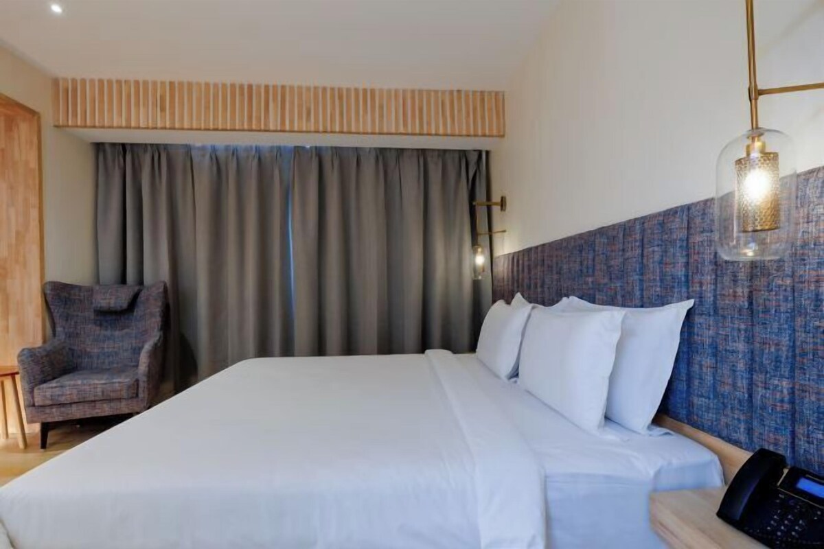 A hotel room with a large bed, a chair, a bedside table with a phone, and a window with curtains.