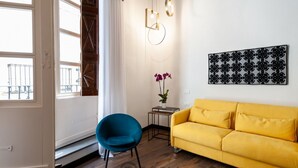 Een kamer met een gele bank, een blauwe stoel, een kleine tafel met een plant en een aan de muur bevestigd kunstwerk.