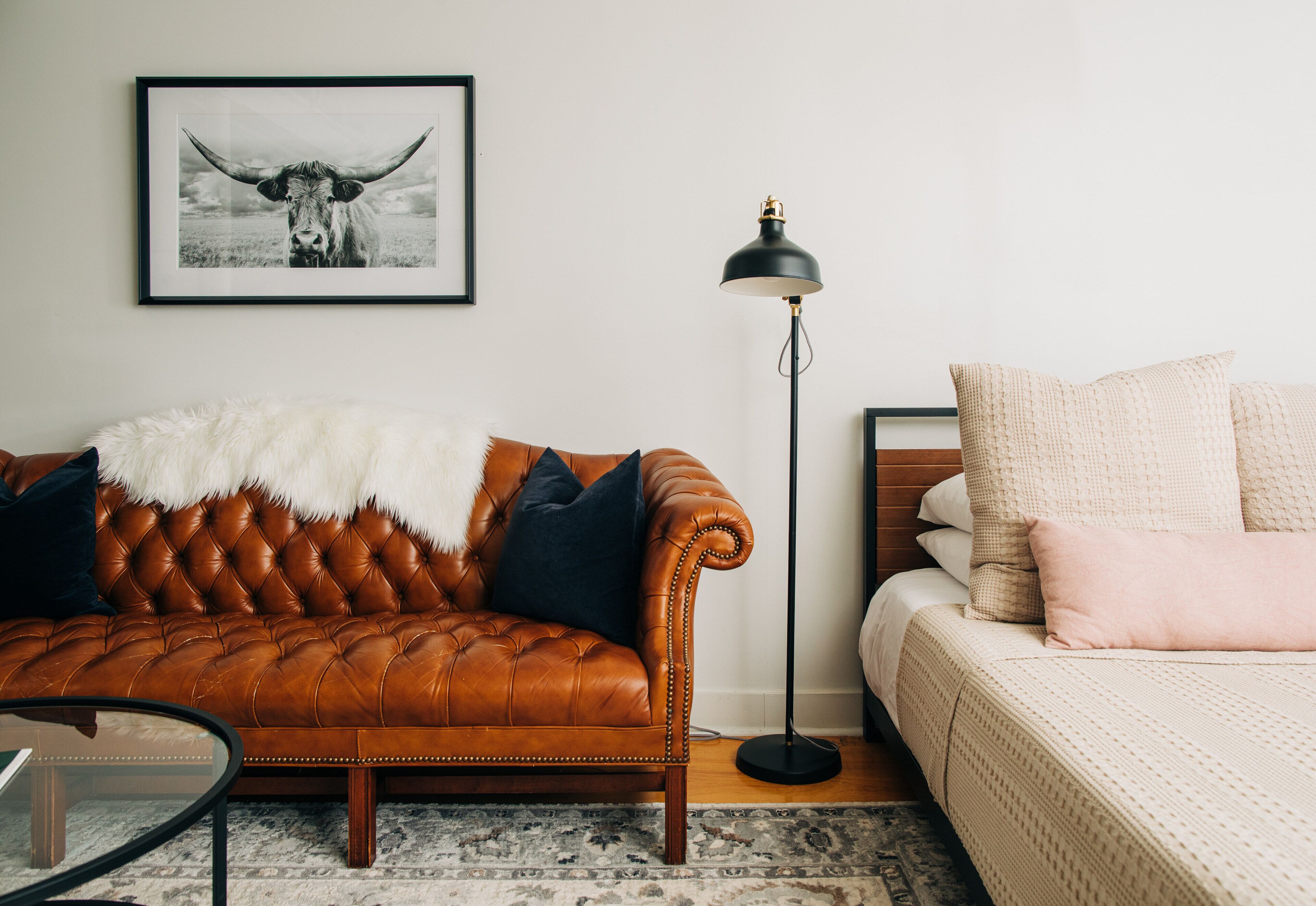 Een kamer met een bruine leren bank, een witte bontmantel, een zwarte vloerlamp en een bed met roze kussens.