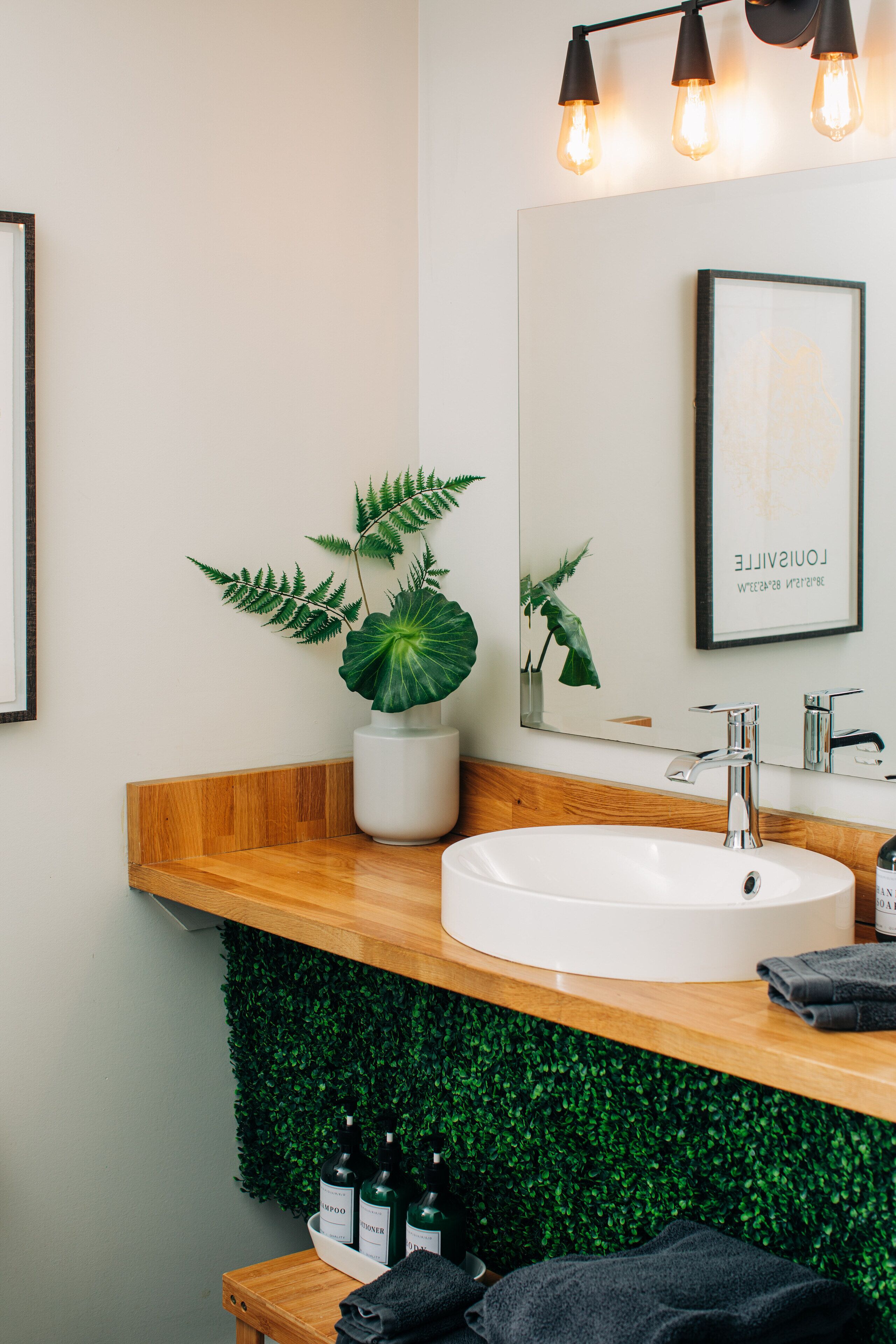 Une salle de bain moderne avec un meuble-lavabo en bois, un lavabo blanc et un miroir. On peut voir une plante en pot sur le meuble-lavabo et une œuvre d’art encadrée au-dessus du miroir.