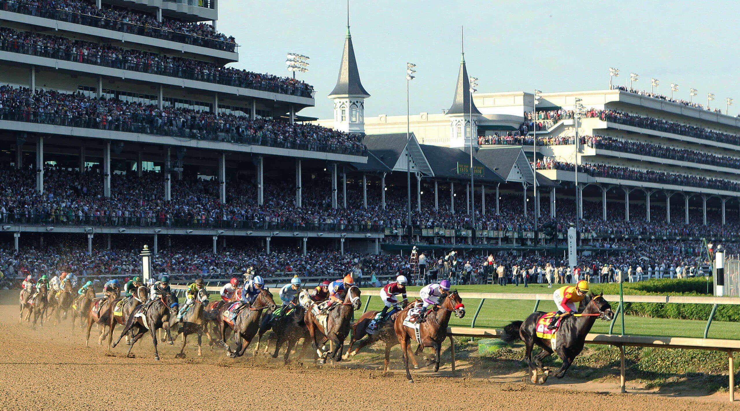 A horse race with multiple horses and jockeys, a large crowd, and a stadium with tiered seating and a clock tower.