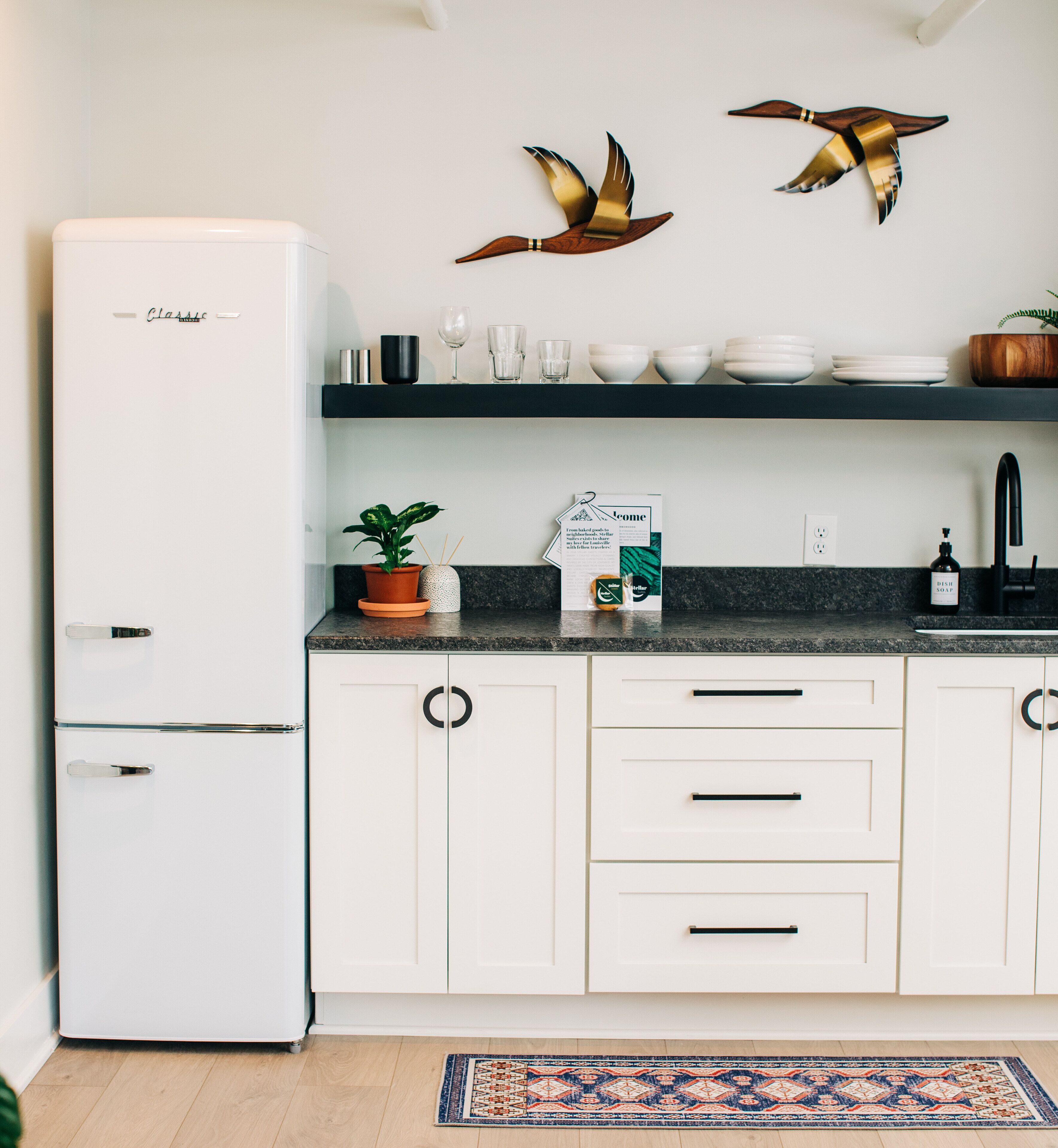 A modern kitchen with white cabinets, a refrigerator, a countertop with a potted plant, and decorative wall art.