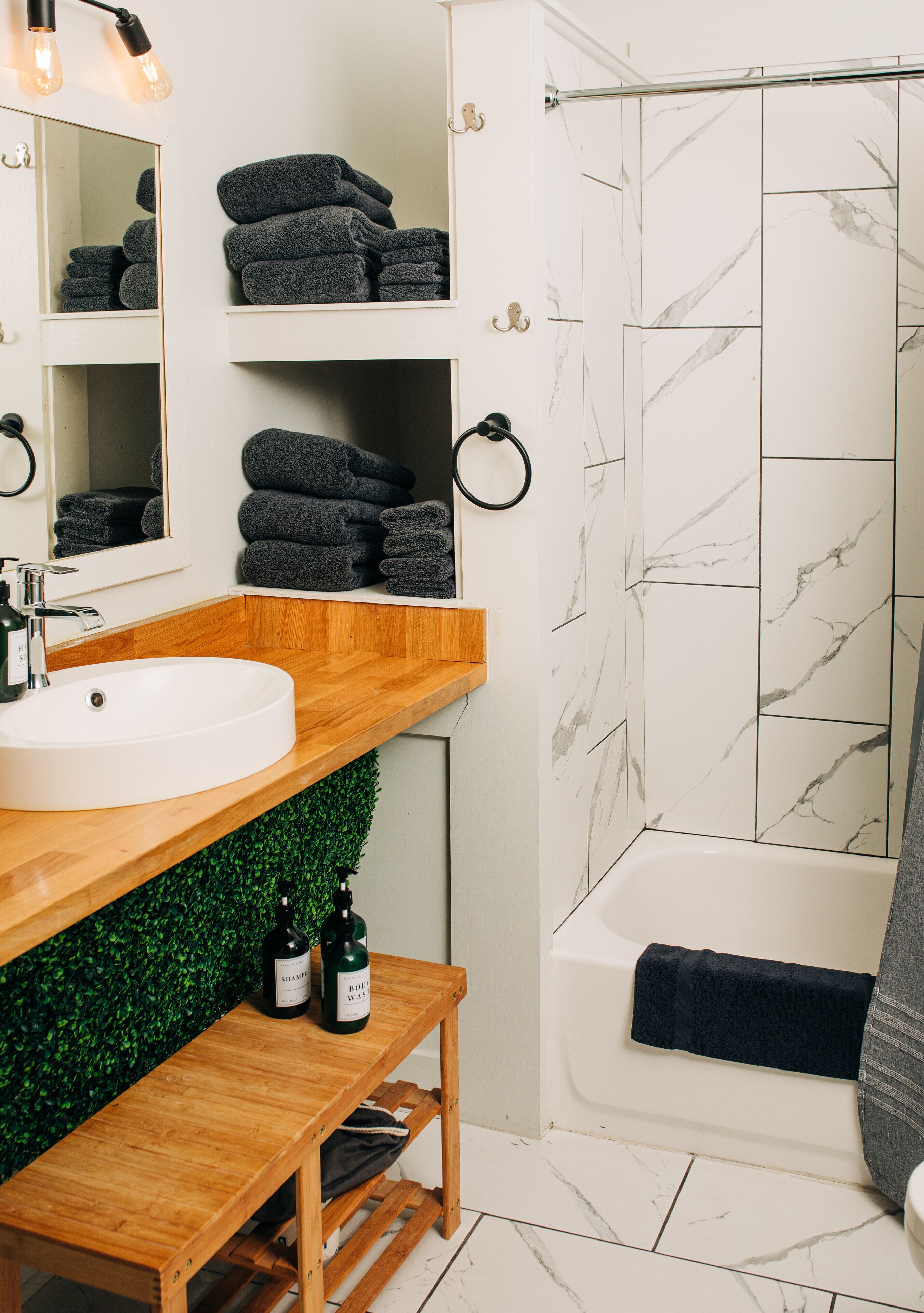Un baño con lavamanos blanco, tocador de madera, pared verde y ducha con azulejos de mármol blanco.