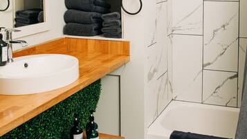 Un baño con lavamanos blanco, tocador de madera, pared verde y ducha con azulejos de mármol blanco.